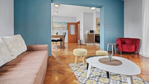 A modern living room with a sofa, ottomans, a coffee table, and a red armchair. A kitchen is visible in the background with a microwave and cabinets. - Seafront Apt, Tastefully Furnished - Neda Smilje (Kampor)