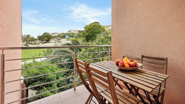 A balcony with a table and chairs, a bowl of fruit, and a view of a green landscape and buildings in the distance. - Seafront Apt, Tastefully Furnished - Neda Smilje (Kampor)
