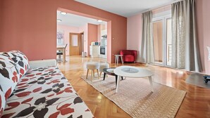 Un salon spacieux avec un canapé, des poufs, une table basse et un fauteuil rouge. On aperçoit une cuisine au fond.