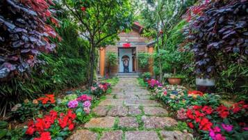A stone pathway leading to a building with a door, surrounded by lush greenery and flowering plants.