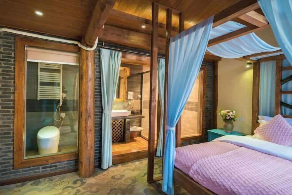A bedroom with a four-poster bed, a window with curtains, a wooden headboard, and a bedside table with a vase of flowers.