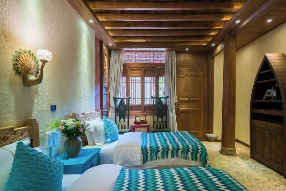 A room with two beds, a wooden ceiling, a decorative wall piece, a window with curtains, and a bookshelf.