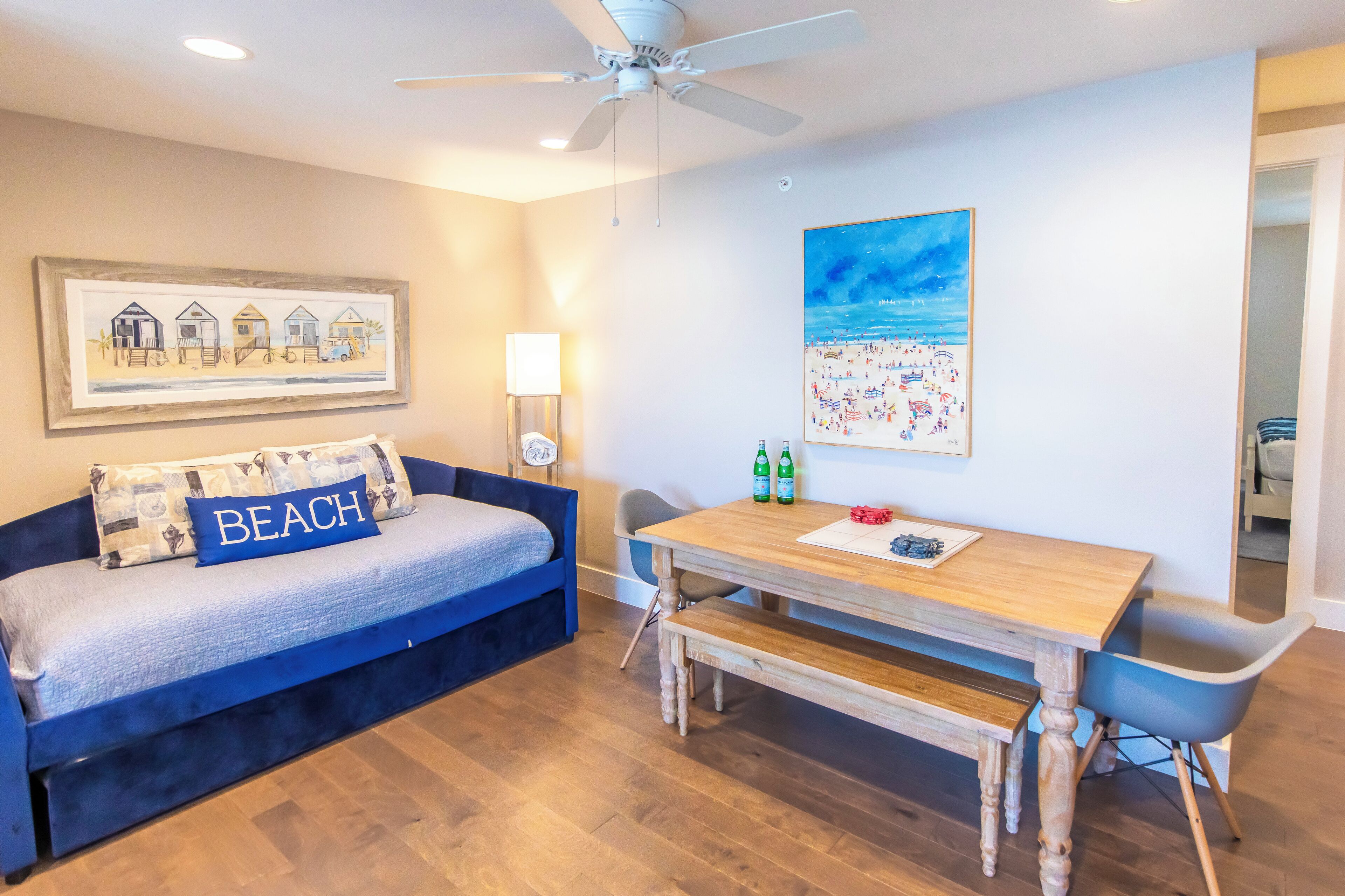 A room with a bed, a table, and a chair. There is a painting on the wall and a ceiling fan.
