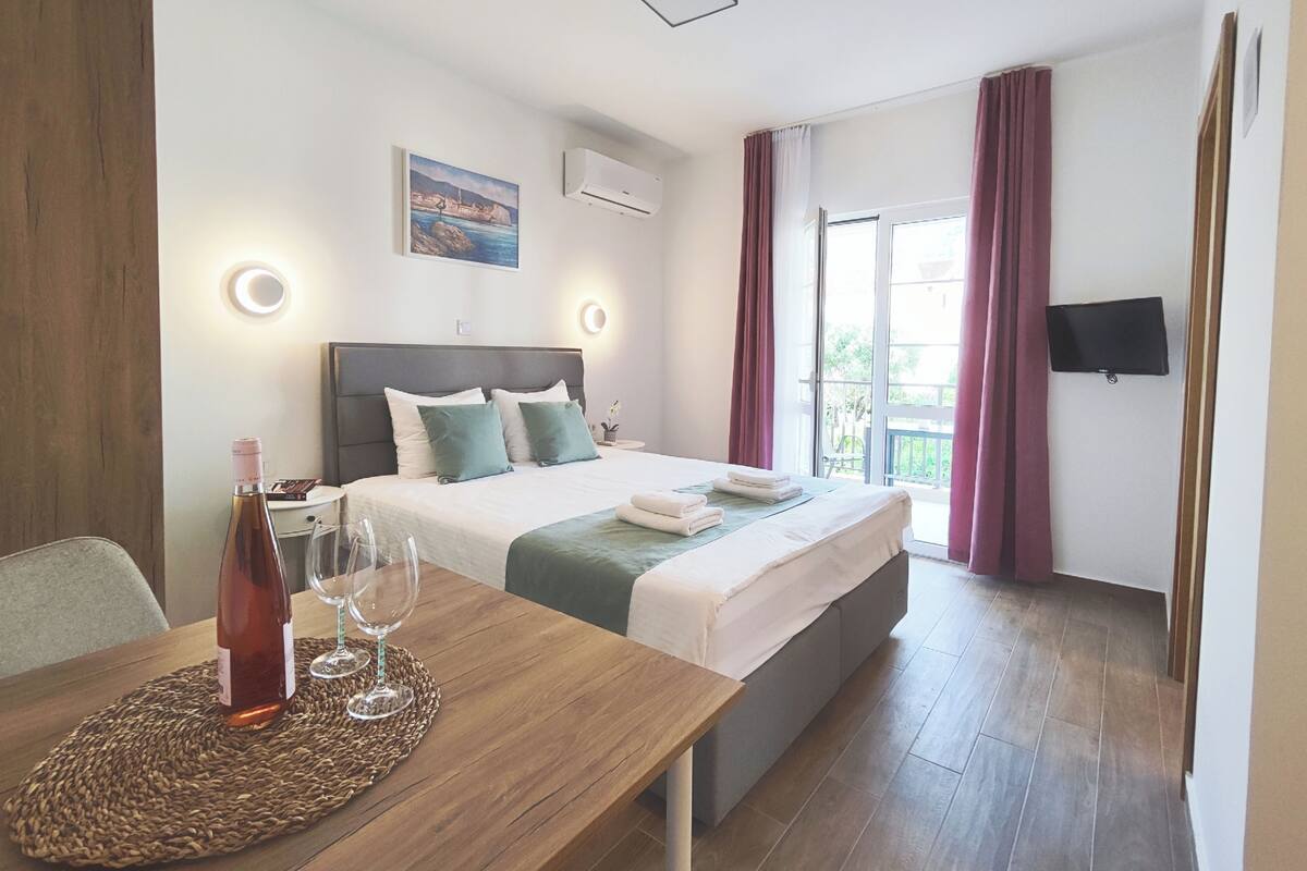 A hotel room with a bed, bedside table with a bottle and glasses, a TV, and a window with curtains.