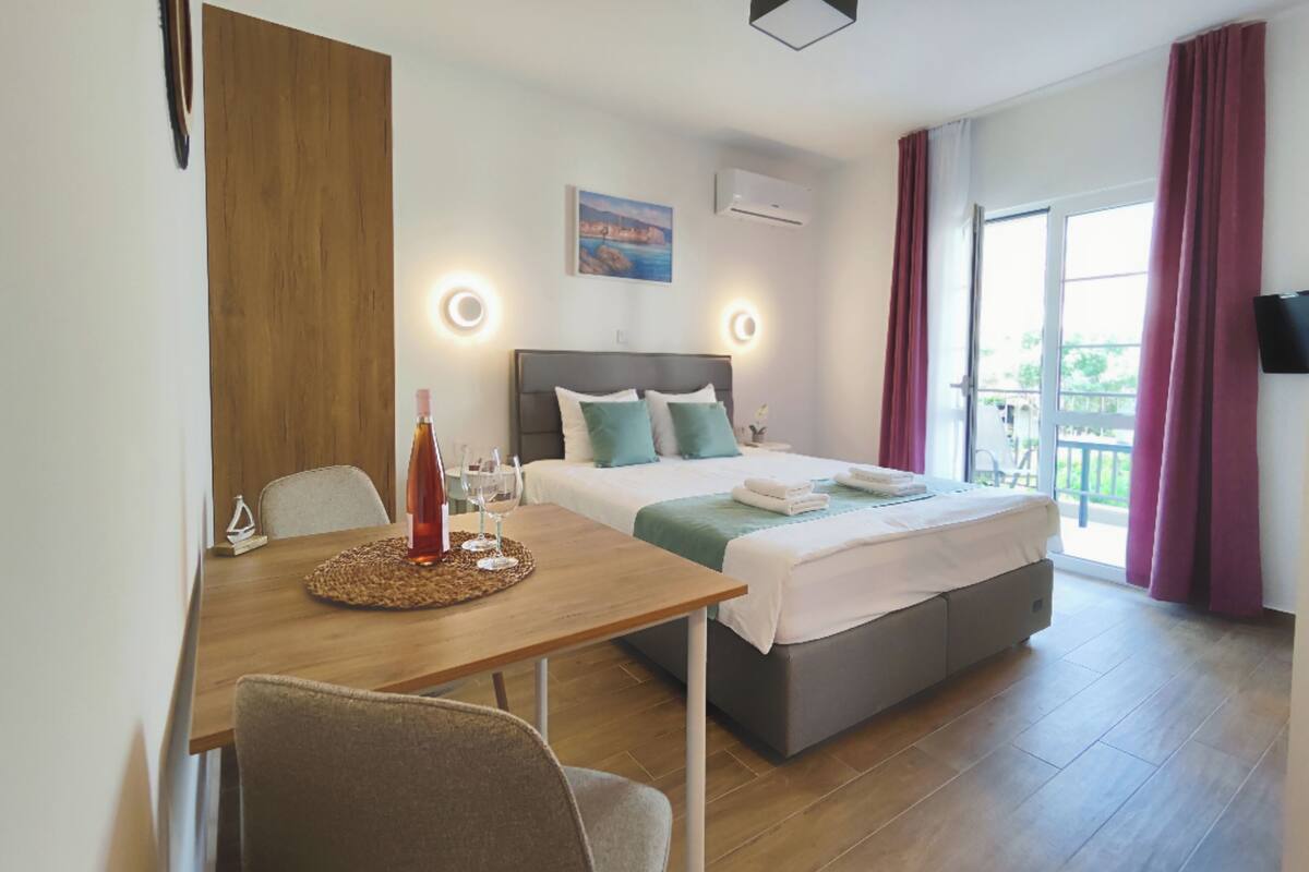A hotel room with a bed, bedside tables, a desk with a chair, a small table with a bottle and glasses, and a TV mounted on the wall.