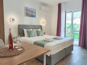 Camera d'albergo con un letto, comodino con vino e bicchieri, un quadro appeso al muro e un balcone con tende.