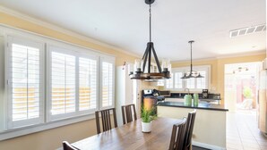 A dining area with a wooden table, chairs, and a potted plant. There is a kitchen in the background with a countertop and appliances.