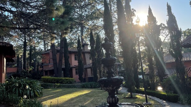 Un jardín con una fuente, rodeado de árboles altos y un edificio con techo de tejas.