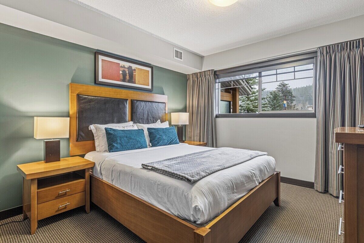 Une chambre d’hôtel bien rangée, avec un grand lit, deux tables de chevet, une petite table, un tableau encadré au mur et une fenêtre avec des rideaux.