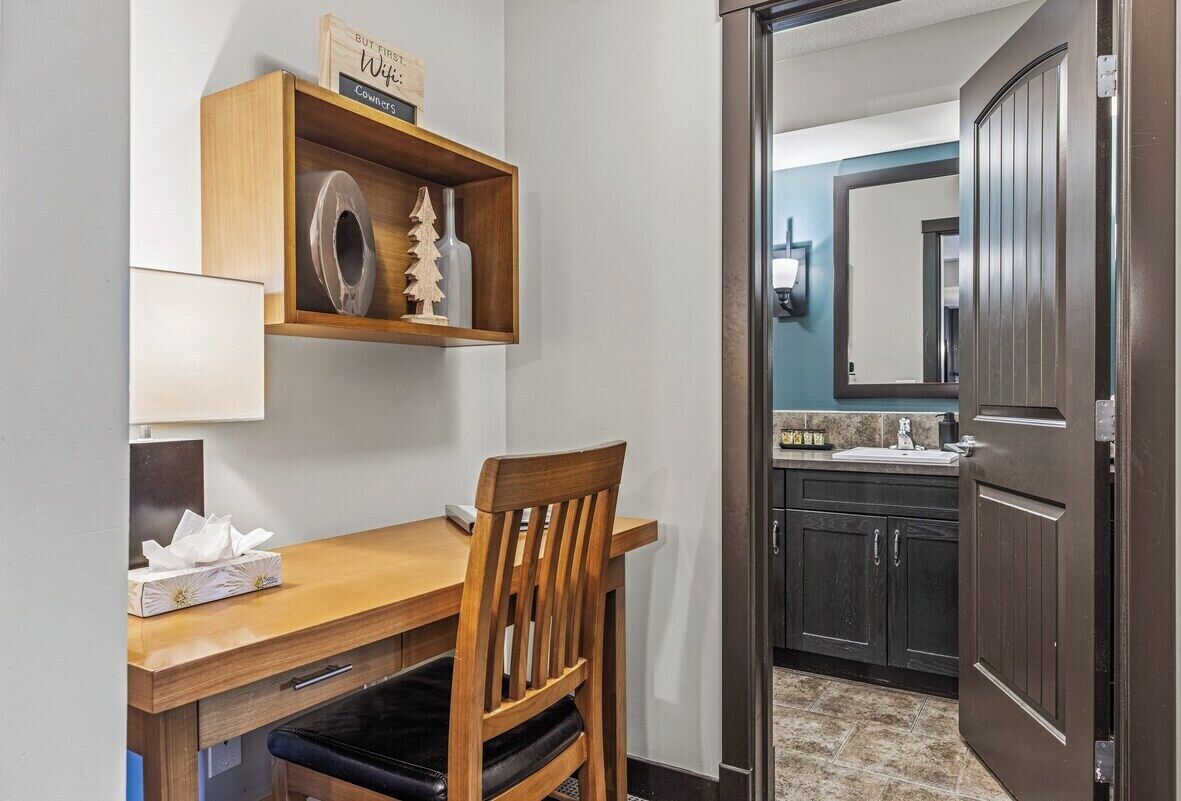 A hotel room with a wooden desk, a chair, a small shelf with decorative items, a mirror, and a dark wooden door.