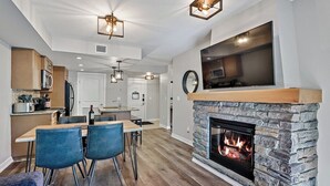 A modern kitchen with a stone fireplace, dining area with chairs, and a large TV mounted above the fireplace.