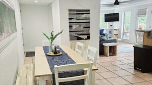 Ein Essbereich mit einem Holztisch, weißen Stühlen und einer blau gemusterten Tischläufer. Auf dem Tisch steht eine Pflanze, und an der Wand ist ein Fernseher angebracht.