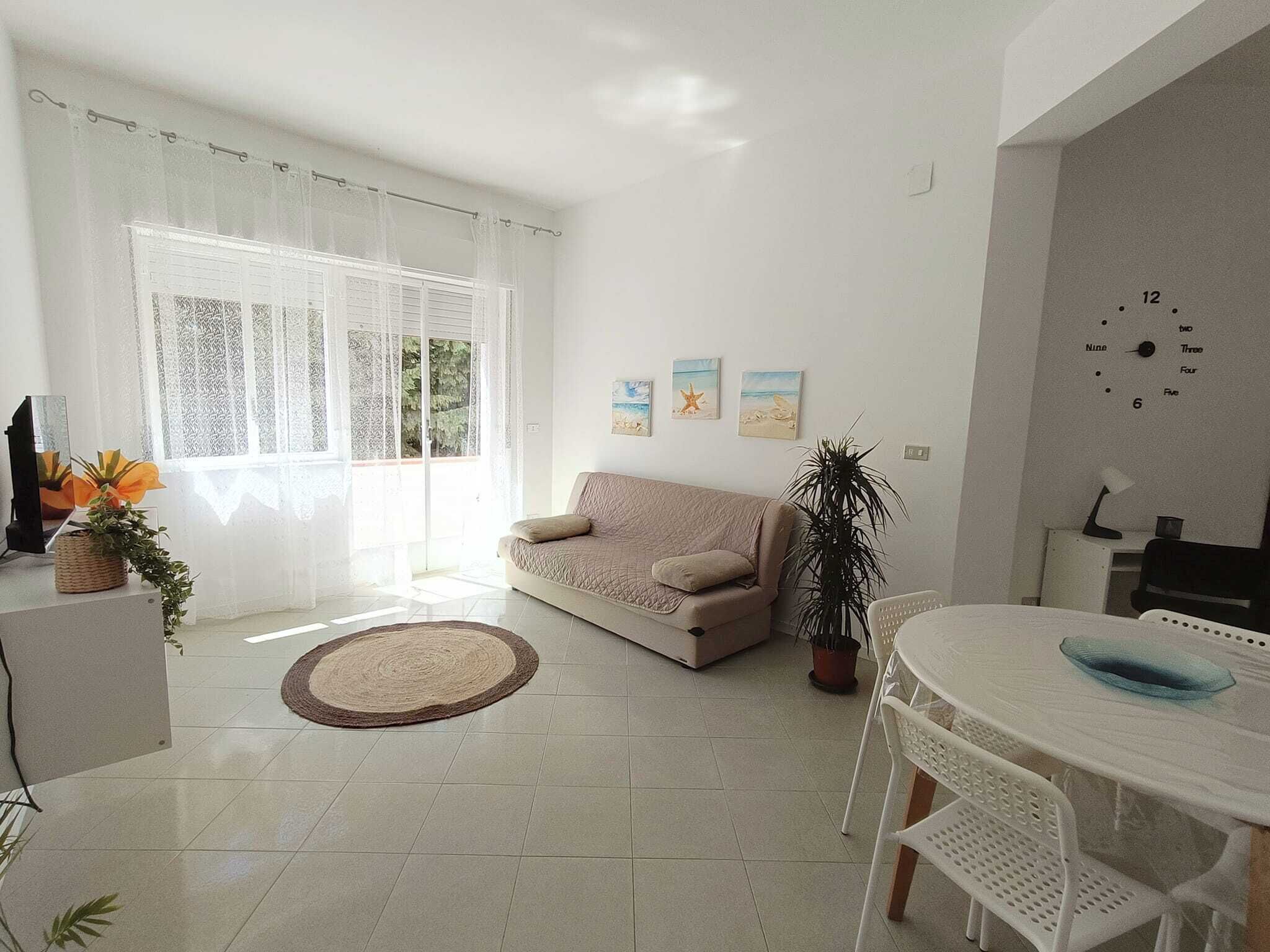 Un salon avec un canapé, une table à manger entourée de chaises, une horloge au mur et une fenêtre avec des rideaux.