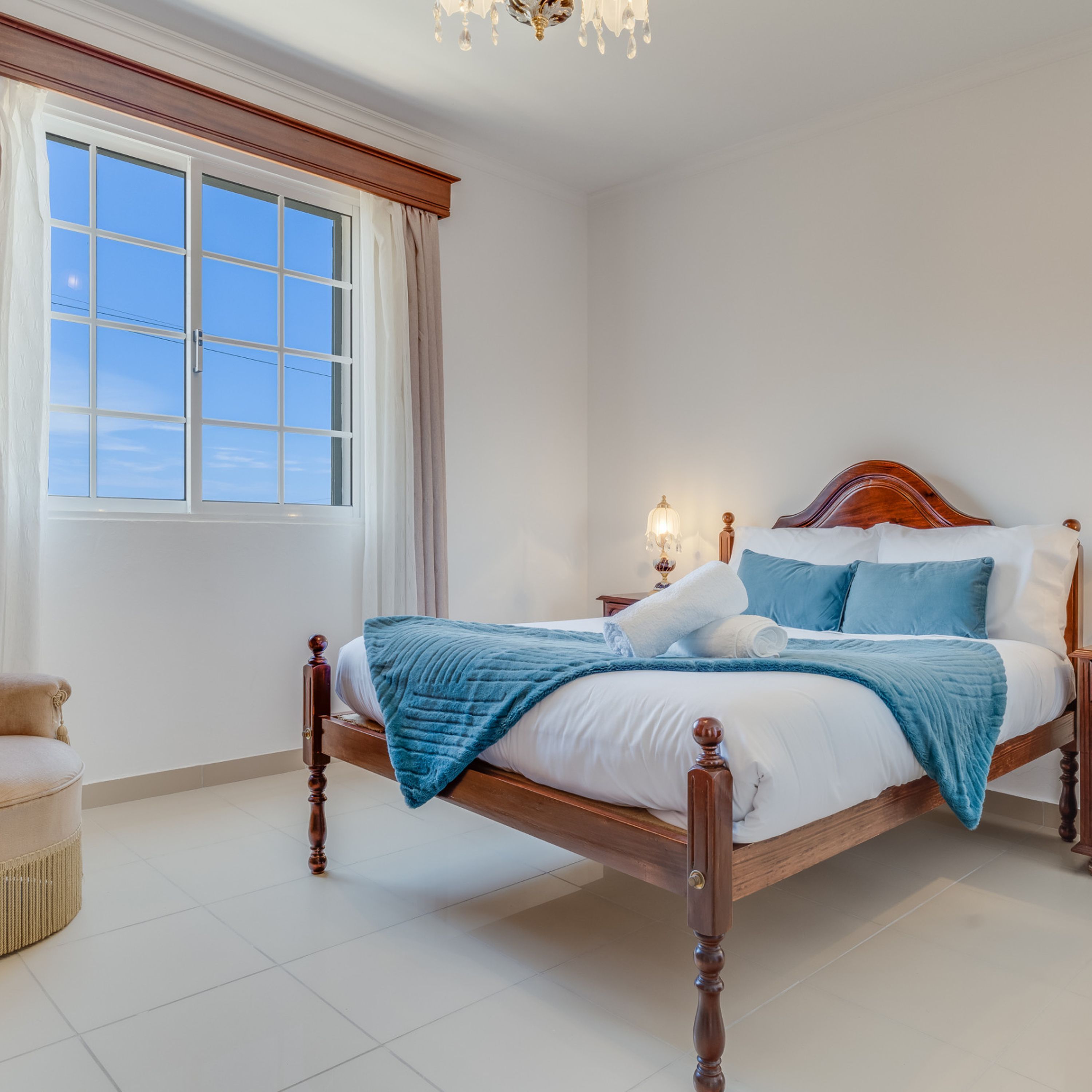 A bedroom with a four-poster bed, a nightstand with a lamp, and a view of the sky through a large window.
