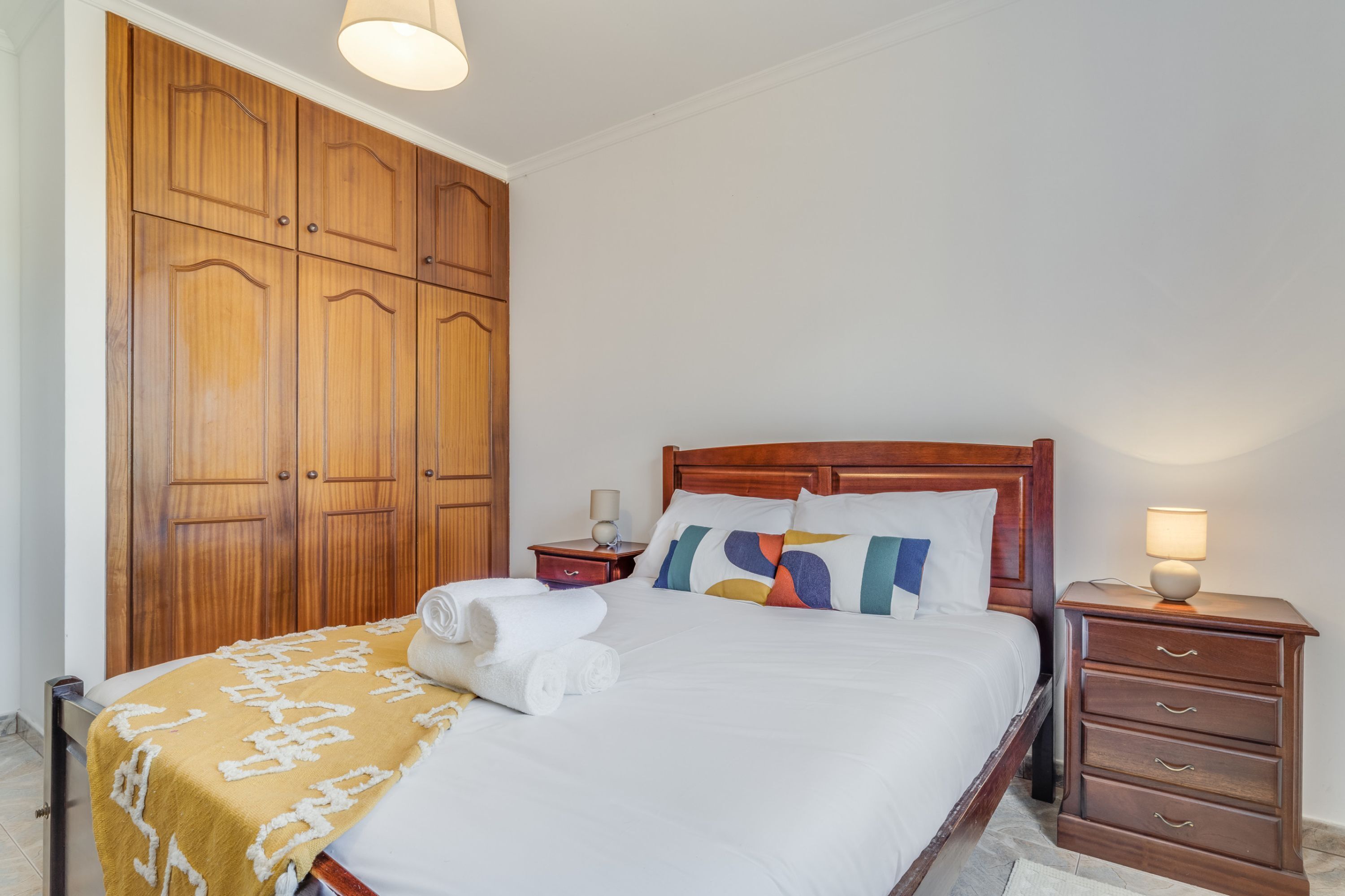 A neatly made bed with a white comforter and a yellow throw blanket, a wooden wardrobe, a bedside table with a lamp, and a nightstand with a lamp.