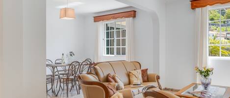 A spacious living room with a beige sofa set, a dining area with a glass table, and a large window offering a view of greenery.