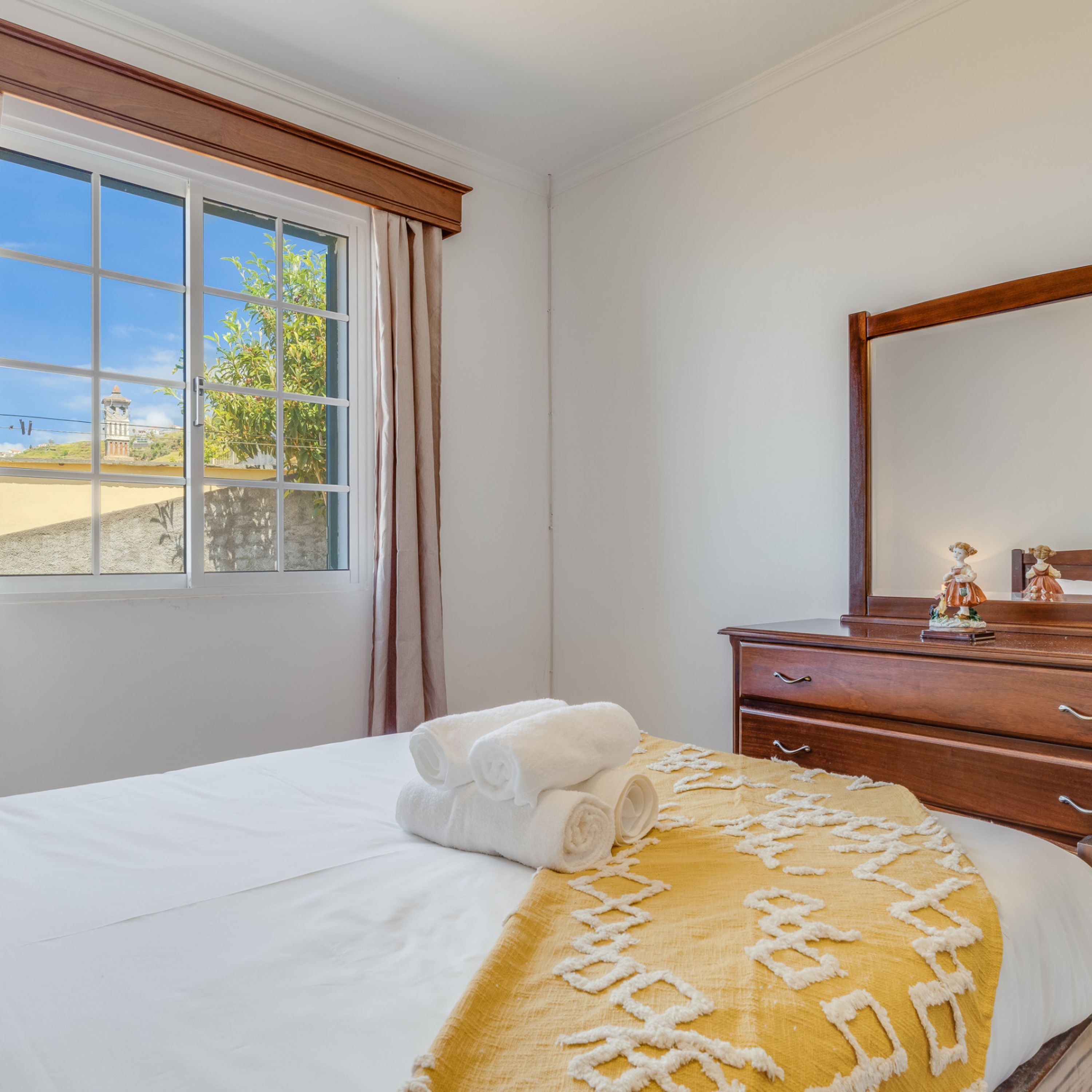 A hotel room with a bed, a dresser, a mirror, and a window with a view of a building and greenery.