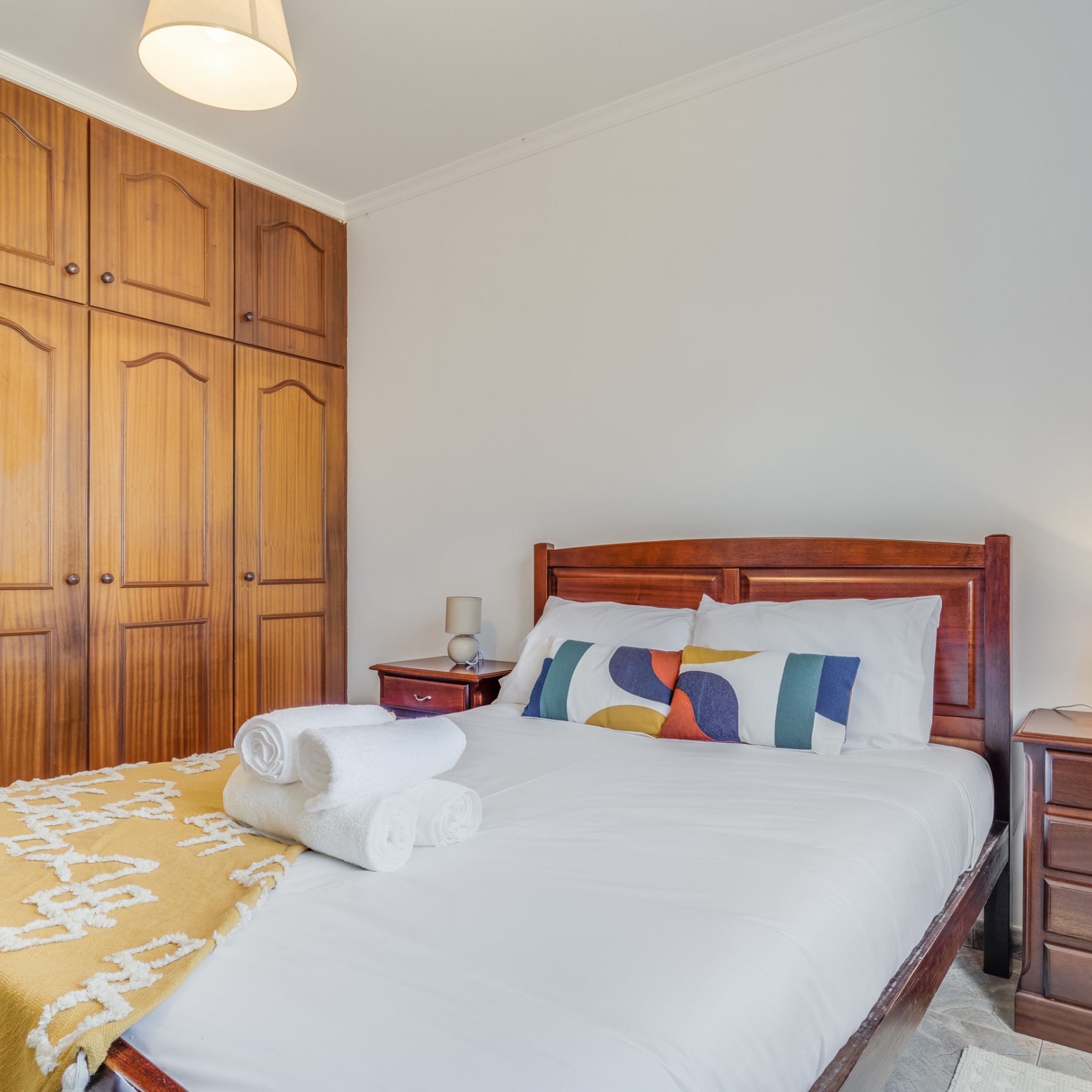 A neatly made bed with white linens and a colorful pillow, a wooden nightstand with a lamp, and a large wooden wardrobe.