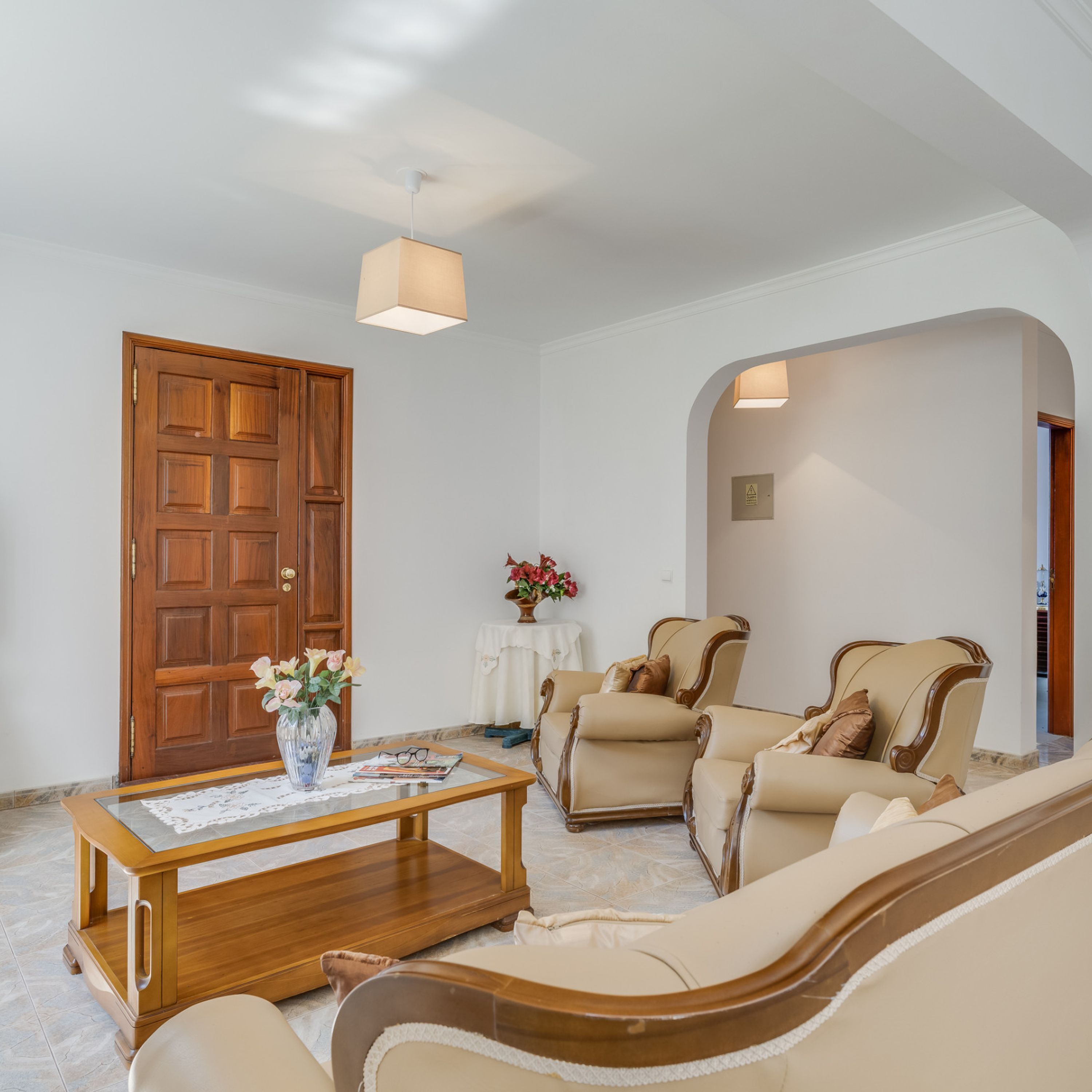 A living room with a wooden door, a coffee table with a vase of flowers, two beige armchairs, and a sofa.