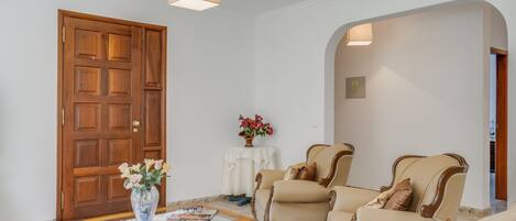 A living room with a wooden door, a coffee table with a vase of flowers, two beige armchairs, and a sofa.
