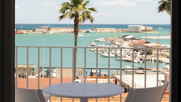 A balcony with two white chairs, a round table, a view of a marina with boats, a palm tree, and a clear blue sky.