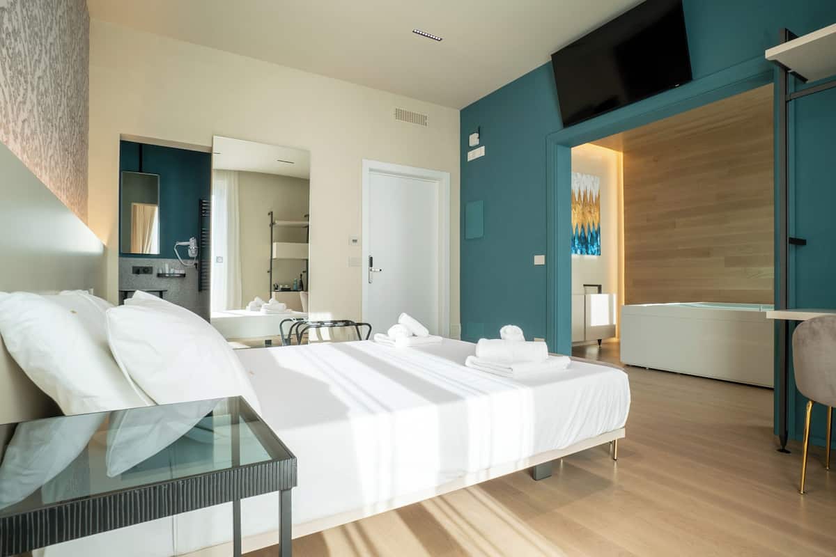 Habitación de hotel moderna con una cama grande, una mesa de vidrio, un baño con ducha y un televisor montado en la pared.