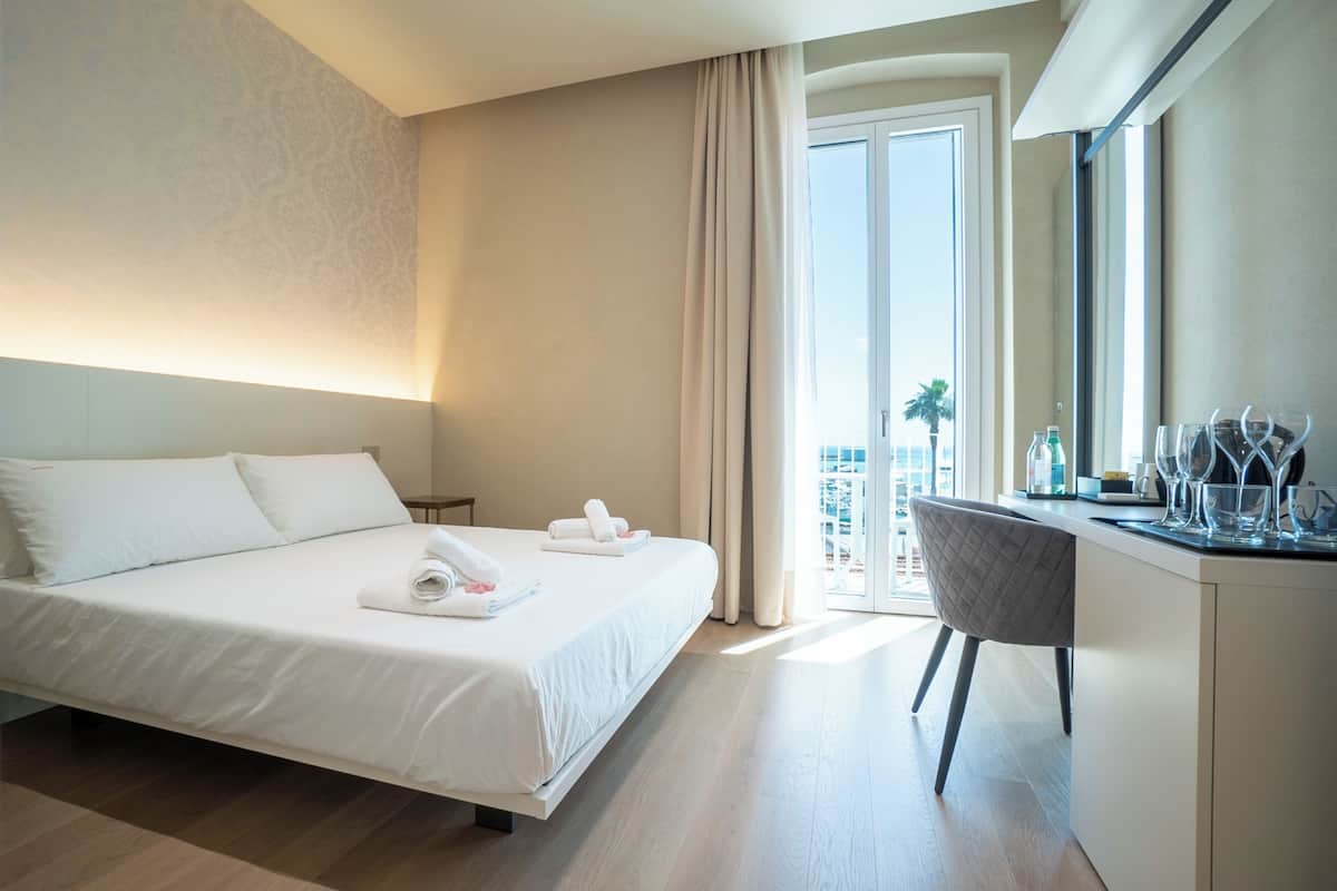 Habitación de hotel con cama, escritorio con silla y ventana grande con vista a palmeras.