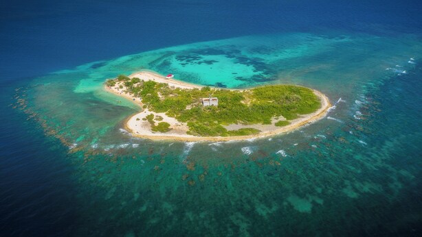 Eine Vogelperspektive zeigt eine kleine Insel mit klarem türkisblauem Wasser, einem Sandstrand und einigen Gebäuden.