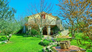 A two-story stone house with a stone staircase leading to the entrance, surrounded by a well-maintained garden with various plants and a small pond.