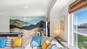 Un soggiorno moderno con un grande quadro raffigurante un paesaggio, un divano con cuscini colorati e vista su una strada attraverso ampie finestre.