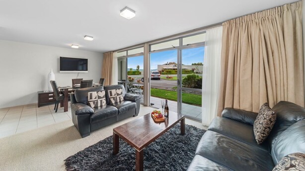 A modern living room with a sofa, coffee table, and TV. There is a dining area with chairs and a table. Large windows offer a view of the outdoors. - 300 Lake Terrace (Taupo)