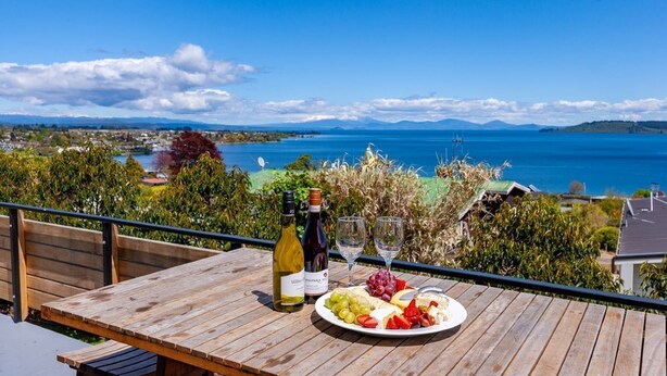 A wooden table with a bottle of wine, a glass, and a plate of fruit and cheese on a balcony overlooking a scenic view of a lake and distant mountains.