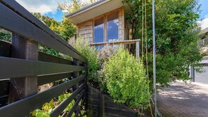 A small wooden house with a window, surrounded by greenery and a metal fence.