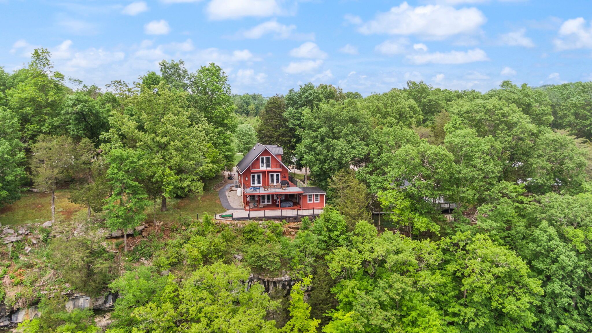 A red house with a large porch, surrounded by dense green forest and a body of water.