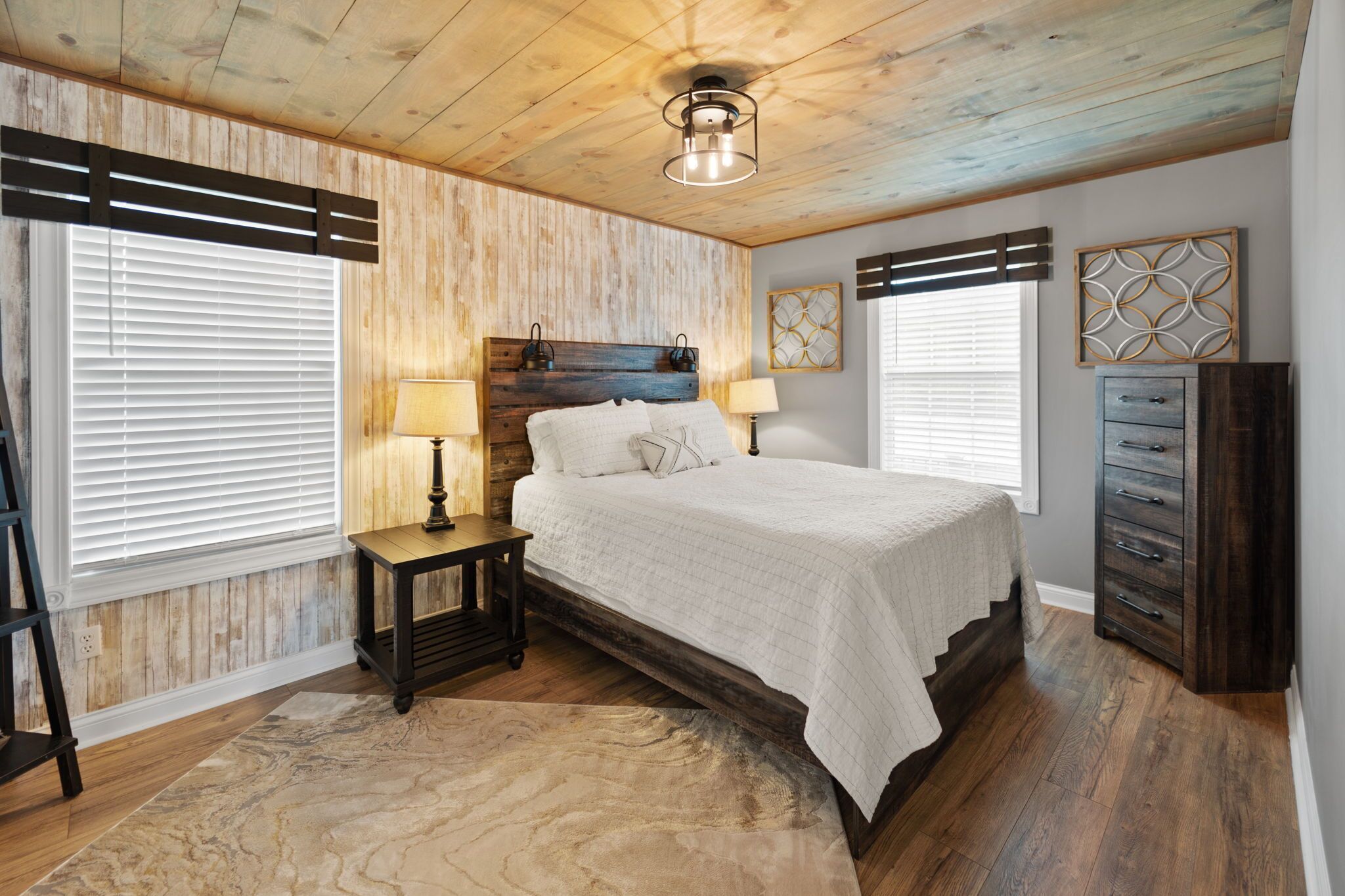 A bedroom with a wooden ceiling, a bed with white bedding, two bedside tables with lamps, and a dresser.