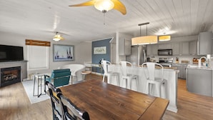 Ein geräumiges Wohnzimmer mit Esstisch aus Holz, weißen Stühlen, Kamin und Deckenventilator.