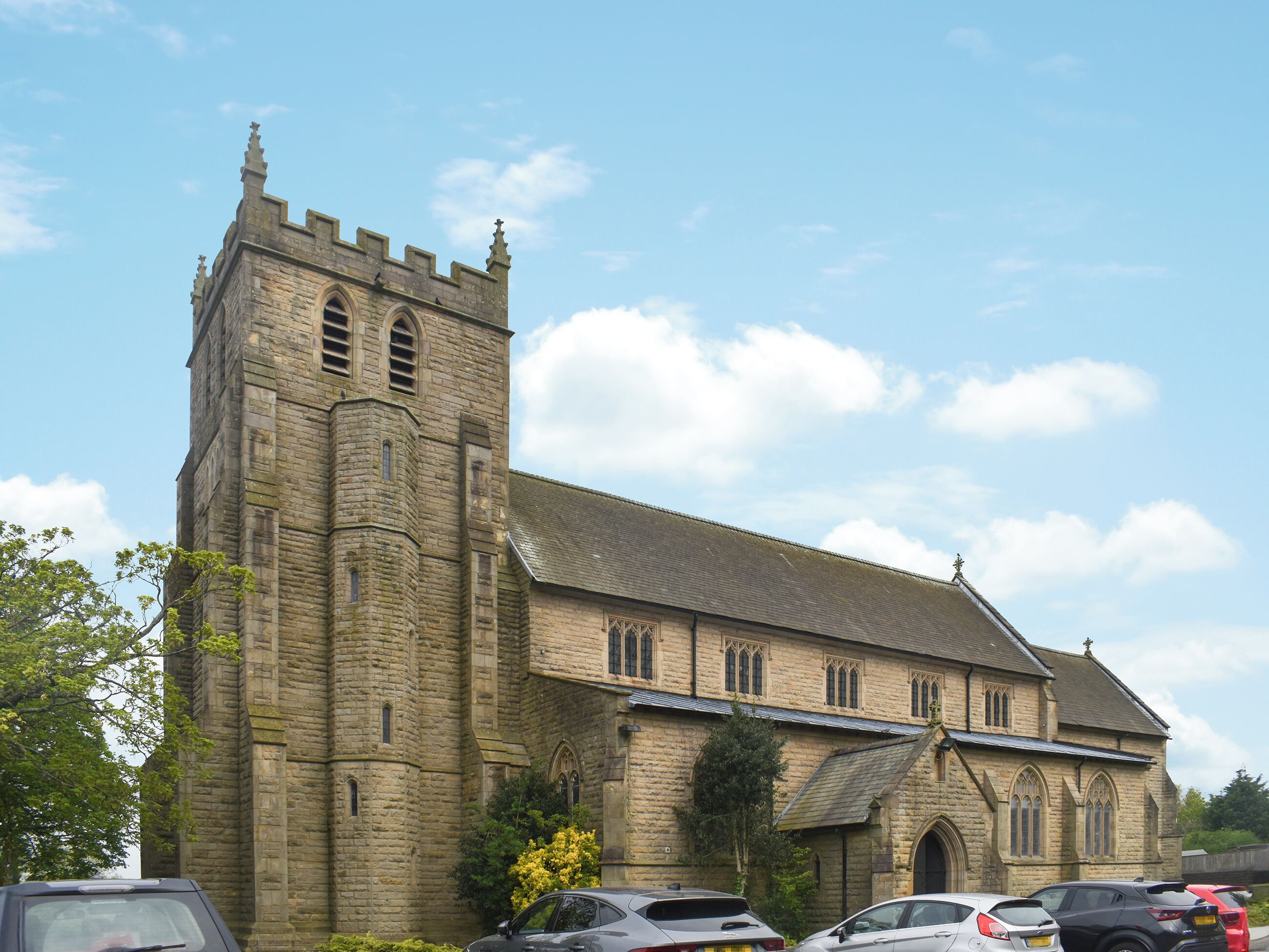 A historic stone building with a tall tower and arched windows, surrounded by parked cars and greenery.