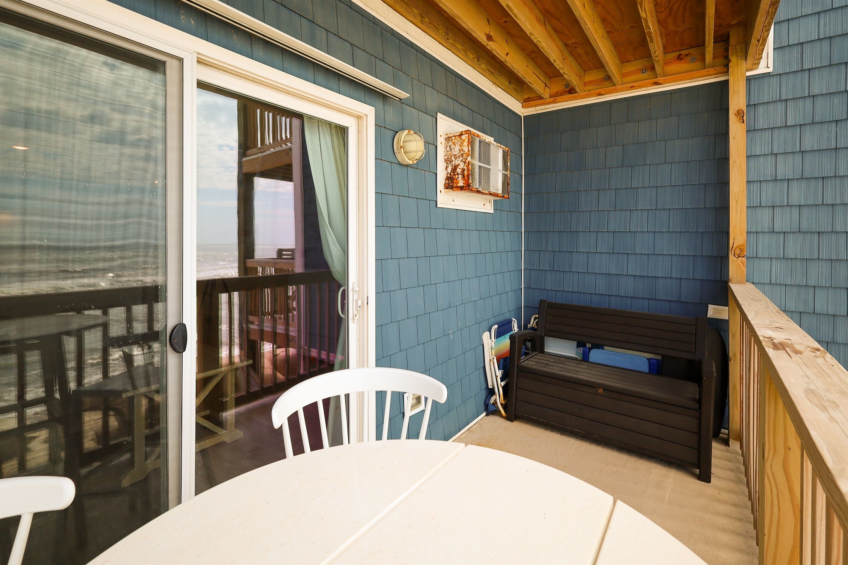 A covered patio area with a table and chairs, a bench, and a view of the ocean through a glass door.
