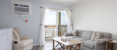 A living room with a sofa, a table, and a chair. There is a window with curtains, a ceiling fan, and a wall-mounted air conditioner.