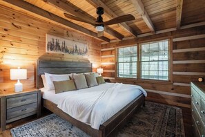 A wooden cabin bedroom with a bed, nightstands, a ceiling fan, and a window. - Gym, Sauna, Spa, Firepit, Game Room + Playground! (Fannin County)