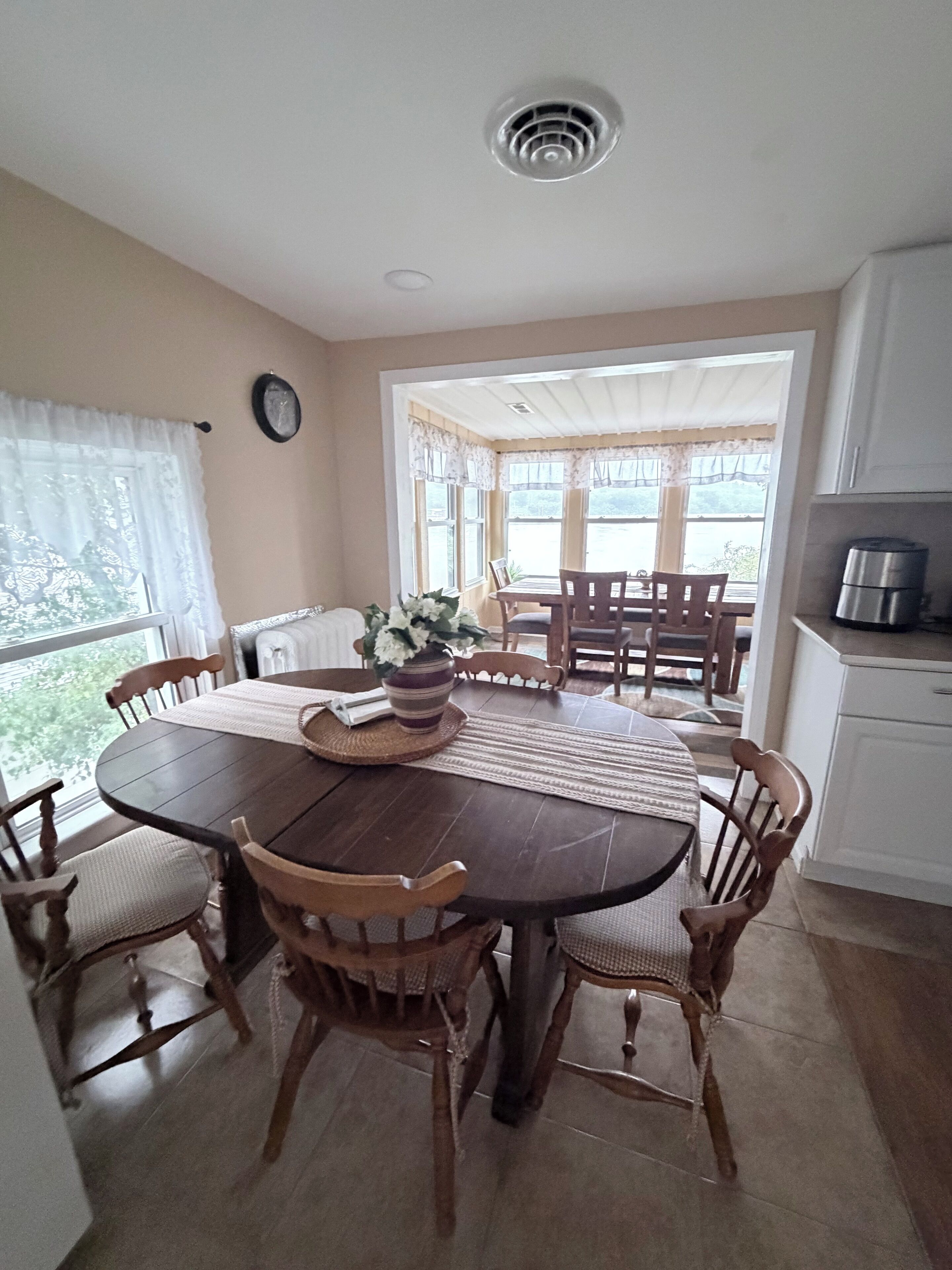 A dining area with a wooden table, chairs, a vase with flowers, and a view through large windows.