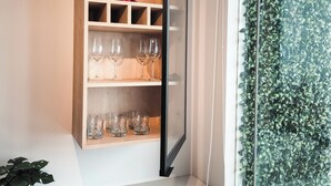 Un interior moderno con una estantería de madera que contiene cristalería y un recipiente rojo, una pequeña planta en maceta y una ventana con un muro cubierto de hojas verdes.