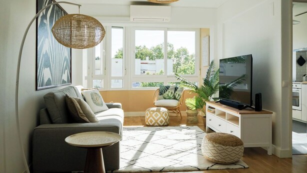 En moderne stue med sofa, sofabord, fjernsyn og et stort vindue med udsigt over grønne områder.