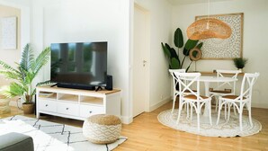 En moderne stue med et fladskærms-tv på et trætv-bord, et spiseområde med hvide stole og et rundt bord, samt en stor stueplante.
