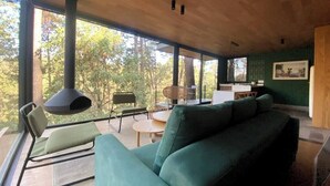 A modern living room with a green sofa, a dining table, and chairs. There is a large window providing a view of trees. - Cabaña Cedro, alojamiento boutique en Avándaro (Valle de Bravo)