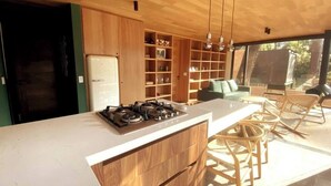 A modern kitchen with a large island, wooden cabinets, and a built-in gas stove. There is a seating area with a sofa and chairs, and a large window providing natural light. - Cabaña Cedro, alojamiento boutique en Avándaro (Valle de Bravo)