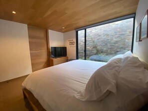 A modern bedroom with a large bed, a TV on a wooden dresser, and a stone wall visible through a large window.