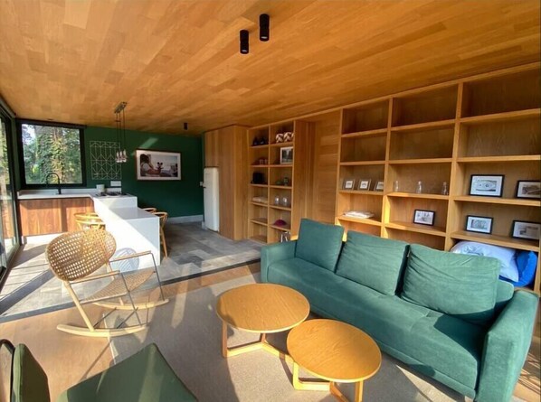 A modern living room with a green sofa, two round wooden tables, and a wooden bookshelf filled with framed pictures.