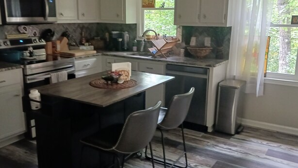 Una cocina con una isla central, dos taburetes de bar, microondas, estufa y varios electrodomésticos.