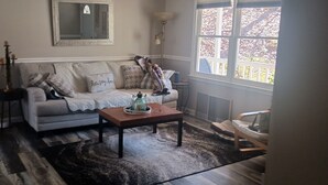 Una sala de estar con un sofá gris, una mesa de centro de madera y un ventanal con vistas a árboles.